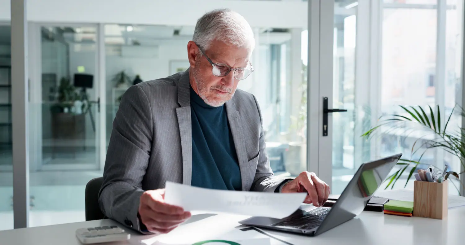 man-reading-paperwork-office-with-laptop-financial-statement-budget-proposal-report-mature-person-glasses-document-with-computer-finance-management-planning-review